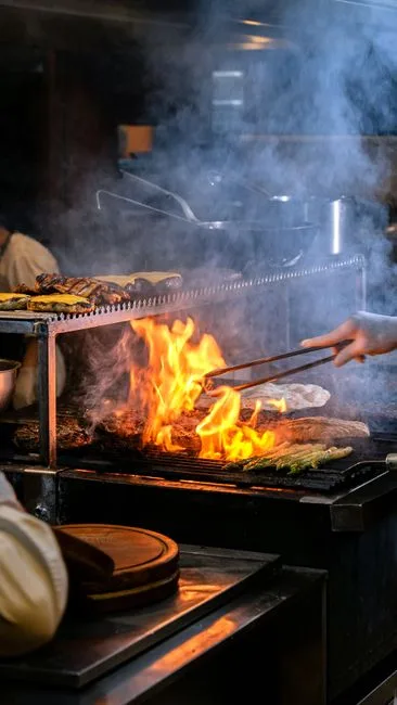 Chef grilling meat with open flames in a commercial kitchen — grease and smoke captured by overhead exhaust hood